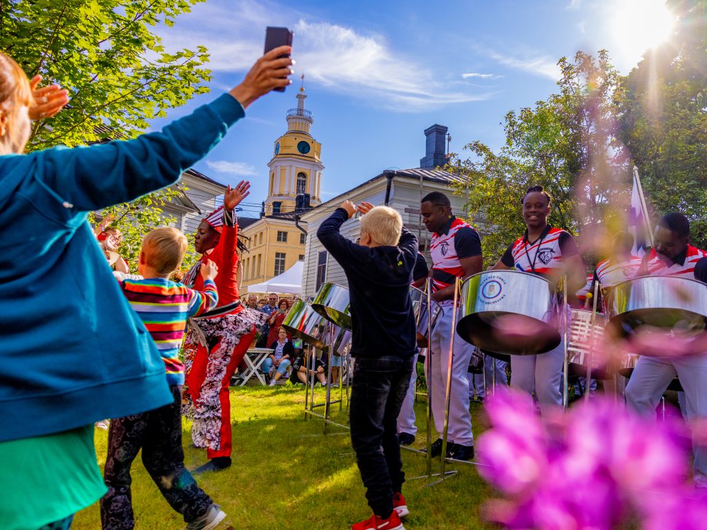 lapset tanssimassa ulkona rumpaleiden soittaessa. Haminan Raatihuone näkyy taustalla.